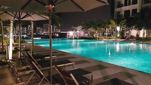 A nighttime pool area with lounge chairs and umbrellas, surrounded by palm trees and a cityscape in the background.
