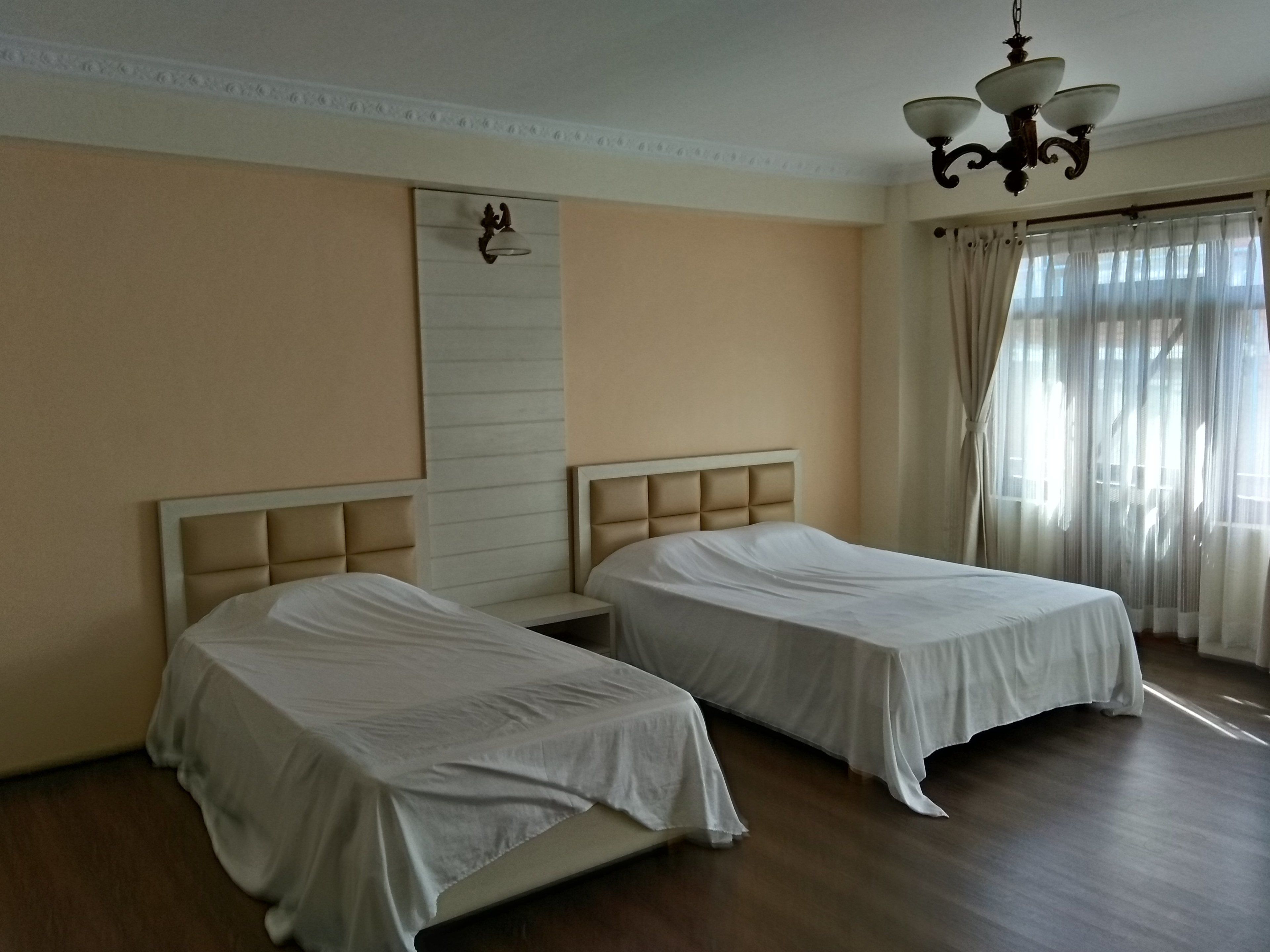 A hotel room with two single beds, a large mirror, a chandelier, and a window with sheer curtains.