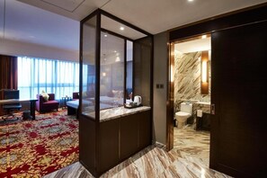A hotel room with a large window, a bed, a desk with a computer, a red carpet, and a bathroom with a marble floor.