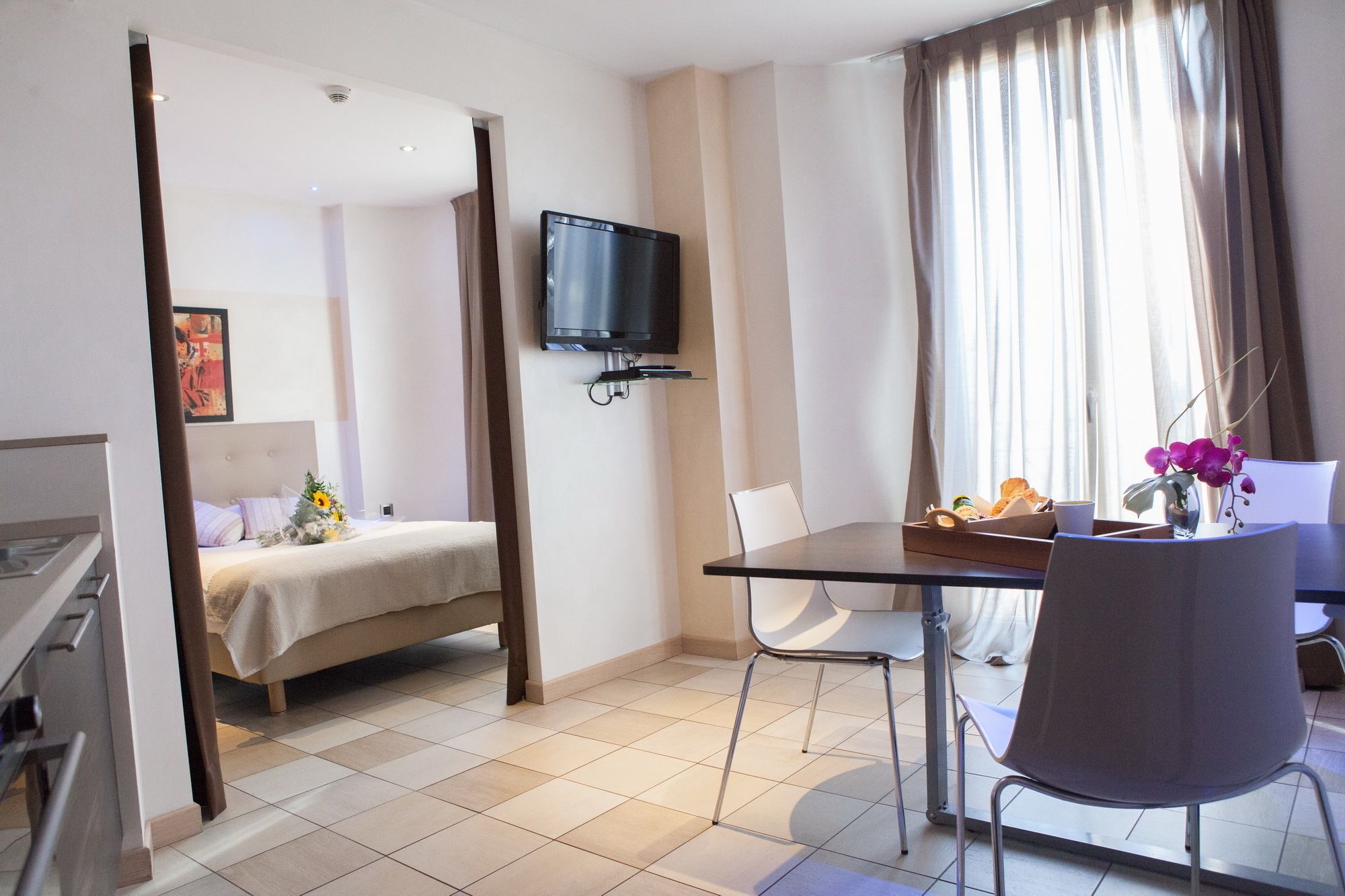 Une chambre d’hôtel comprenant un lit, un téléviseur fixé au mur, une table à manger avec des chaises et une fenêtre avec des rideaux.