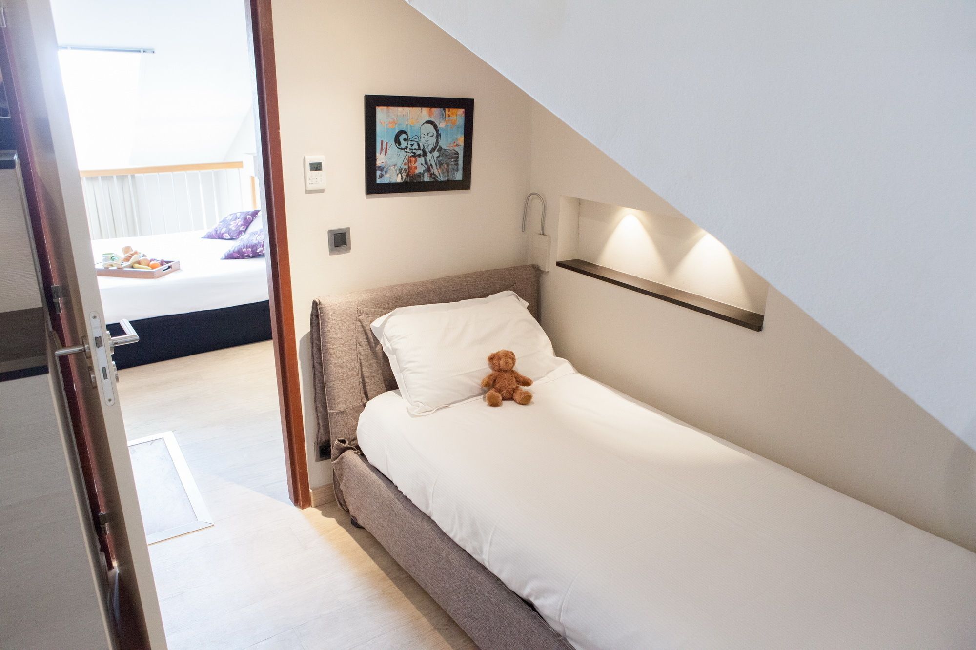 A small, neatly made bed with a teddy bear, a painting on the wall, and a bedside table with a lamp.