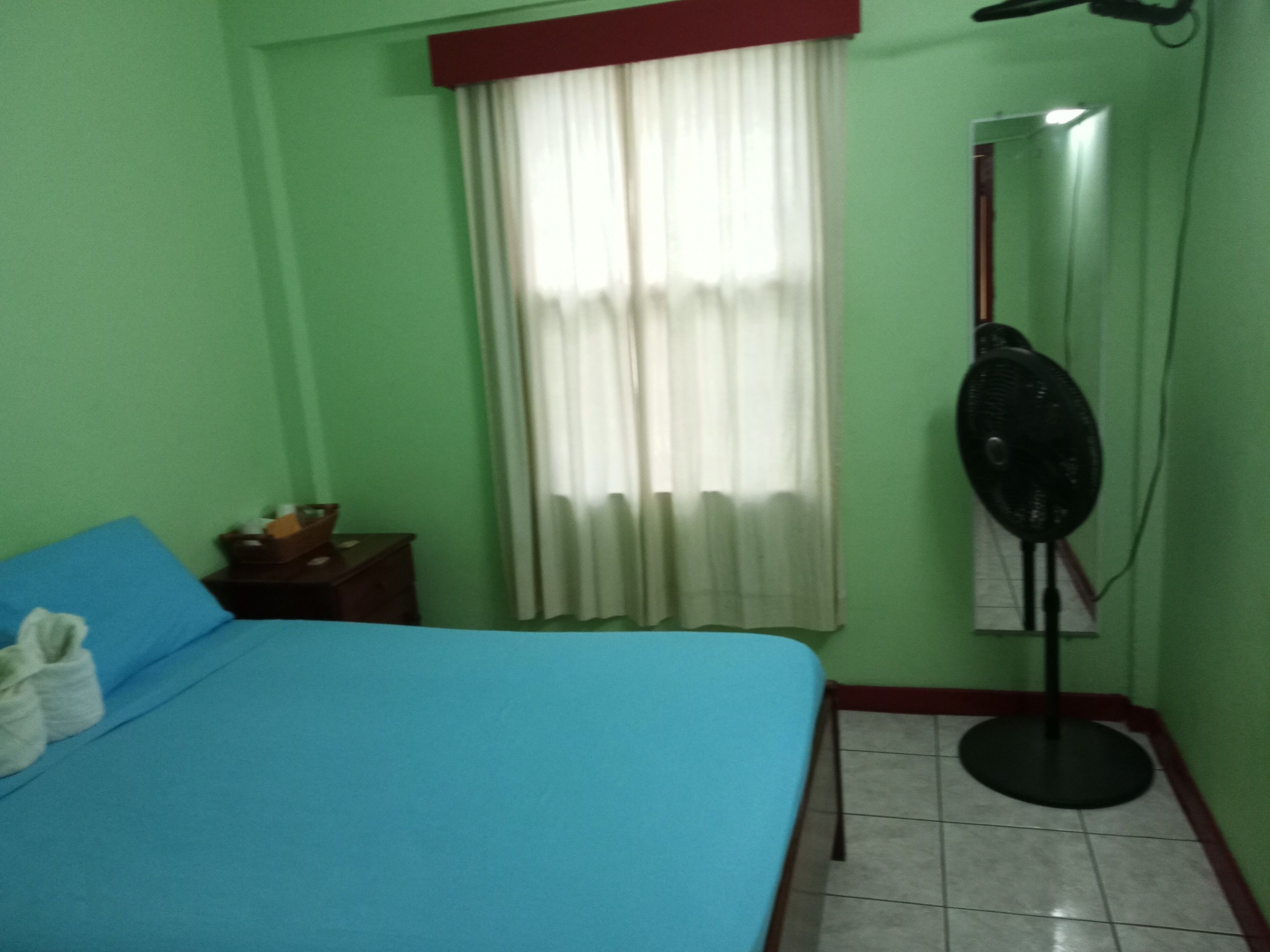 Quarto com cama, uma mesinha, um ventilador e janela com cortinas.