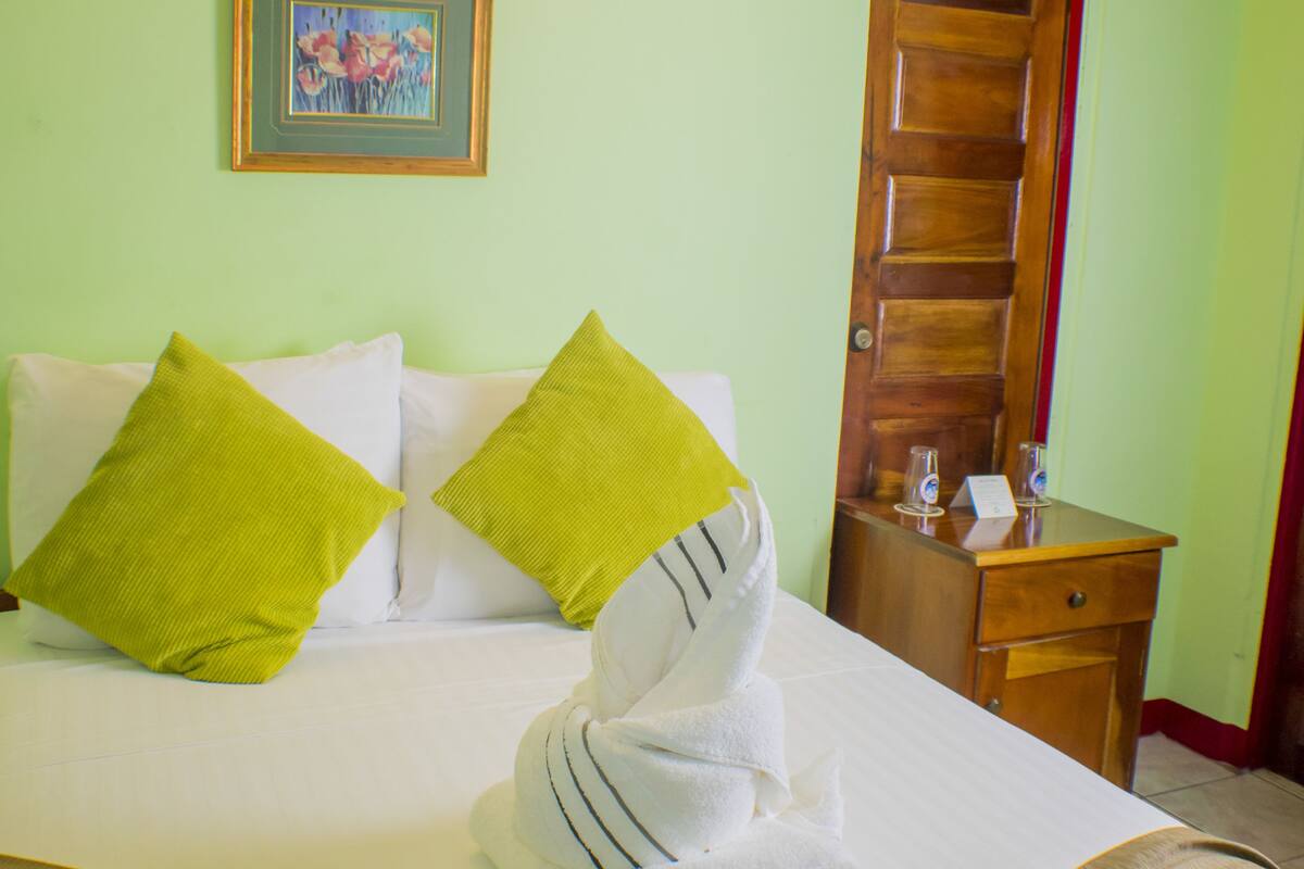 Una cama bien tendida con una manta marrón, dos almohadas verdes y una toalla blanca doblada al lado.