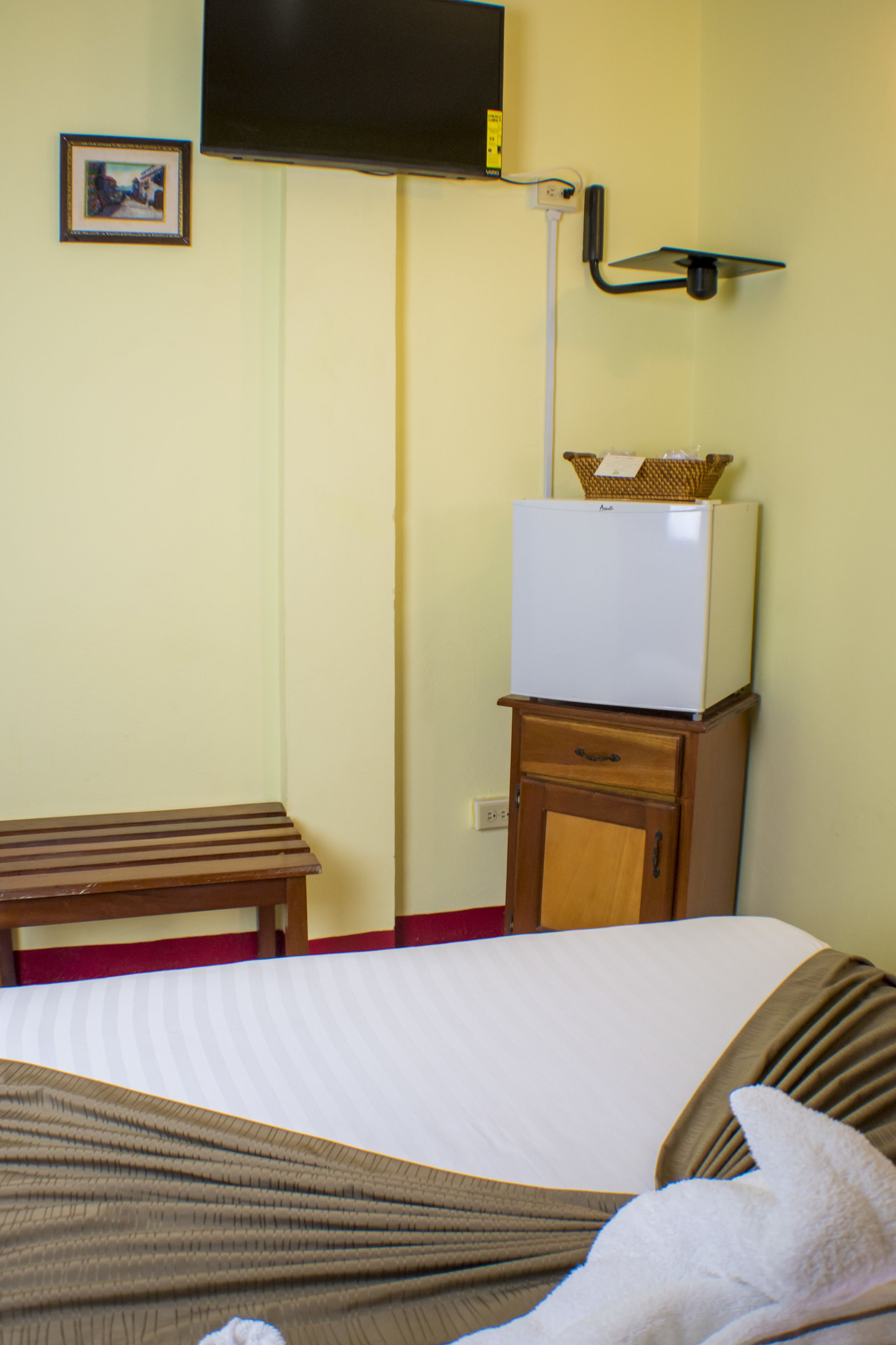 A hotel room with a bed, a small wooden table, a white refrigerator, and a television mounted on the wall.
