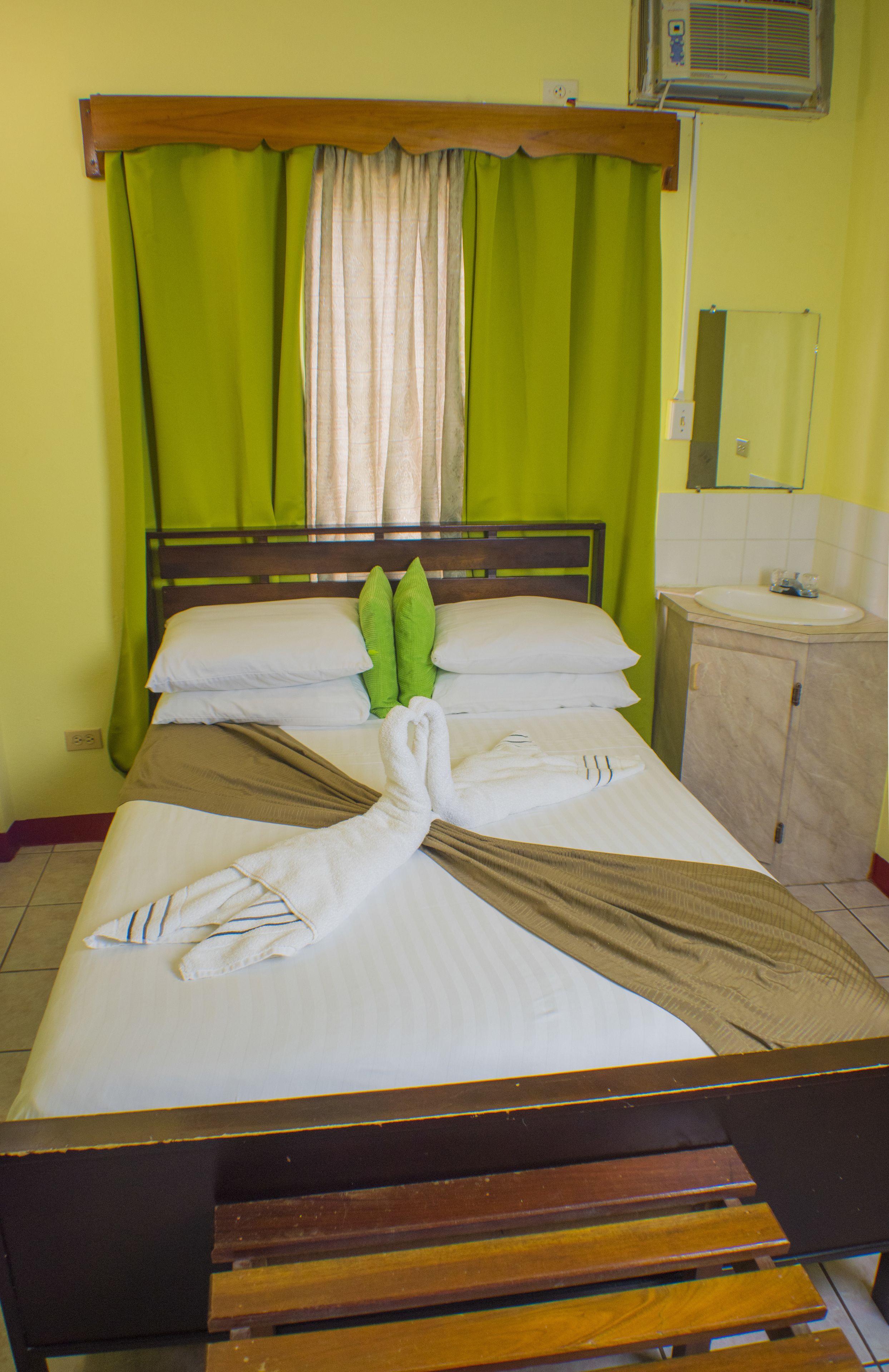 Habitación de hotel con cama, cortinas verdes, cabecero de madera, mesita de noche y baño con lavamanos y espejo.