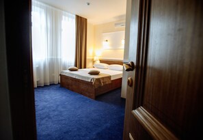 Une chambre d’hôtel avec un grand lit, une tête de lit en bois, une moquette bleue et une fenêtre ornée de rideaux légers.