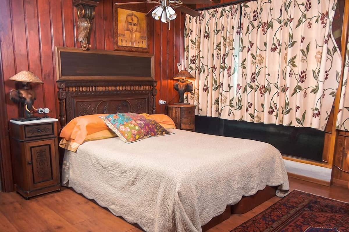 Ein traditionell eingerichtetes Schlafzimmer mit einem hölzernen Kopfteil, einem Bett mit weißer Steppdecke, einem farbenfrohen Kissen, einem Nachttisch mit Lampe und einem Fenster mit blumigem Vorhang.