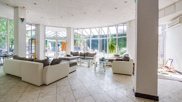 A spacious, well-lit lobby with large windows, a sectional sofa, a coffee table, and a small table with chairs. - CE Napfény Hotel (Siófok)