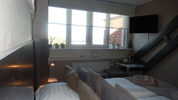 Een slaapkamer met een bed, een bureau, een stoel, een raam met planten en een aan de muur bevestigde televisie.