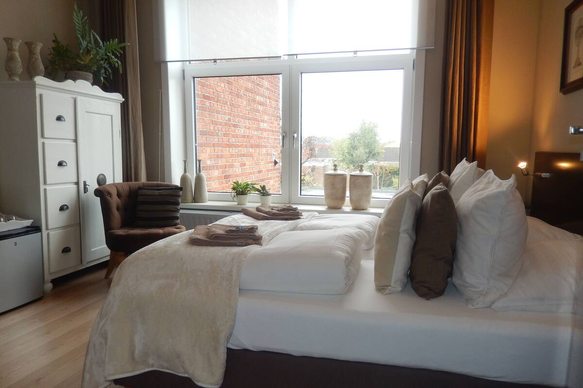 A bedroom with a large bed, a chest of drawers, a chair, and a window with a view of a brick building.