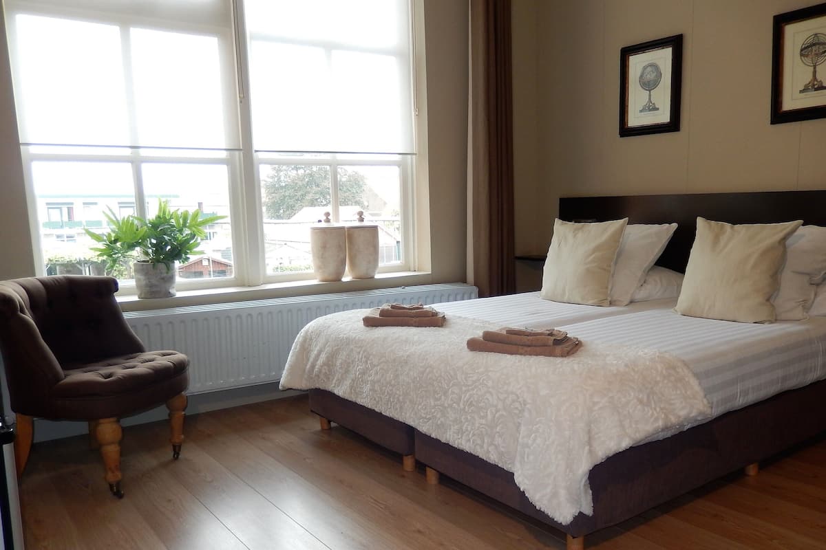 A bedroom with a large bed, a chair, a window with a view, and two potted plants on the windowsill.