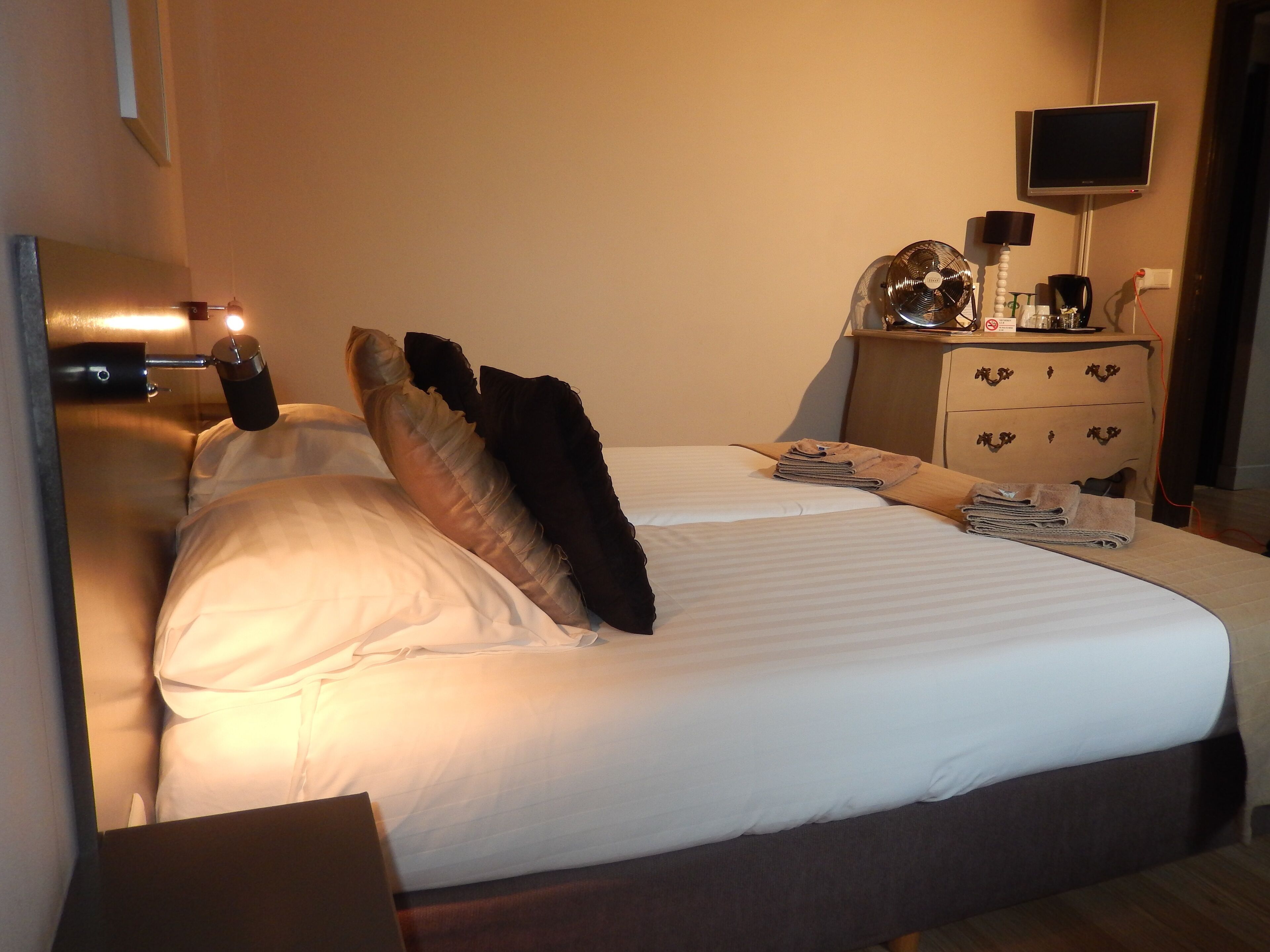 A neatly made bed with white linens and a dark headboard. A wooden nightstand with a lamp and a flat-screen TV is beside the bed.