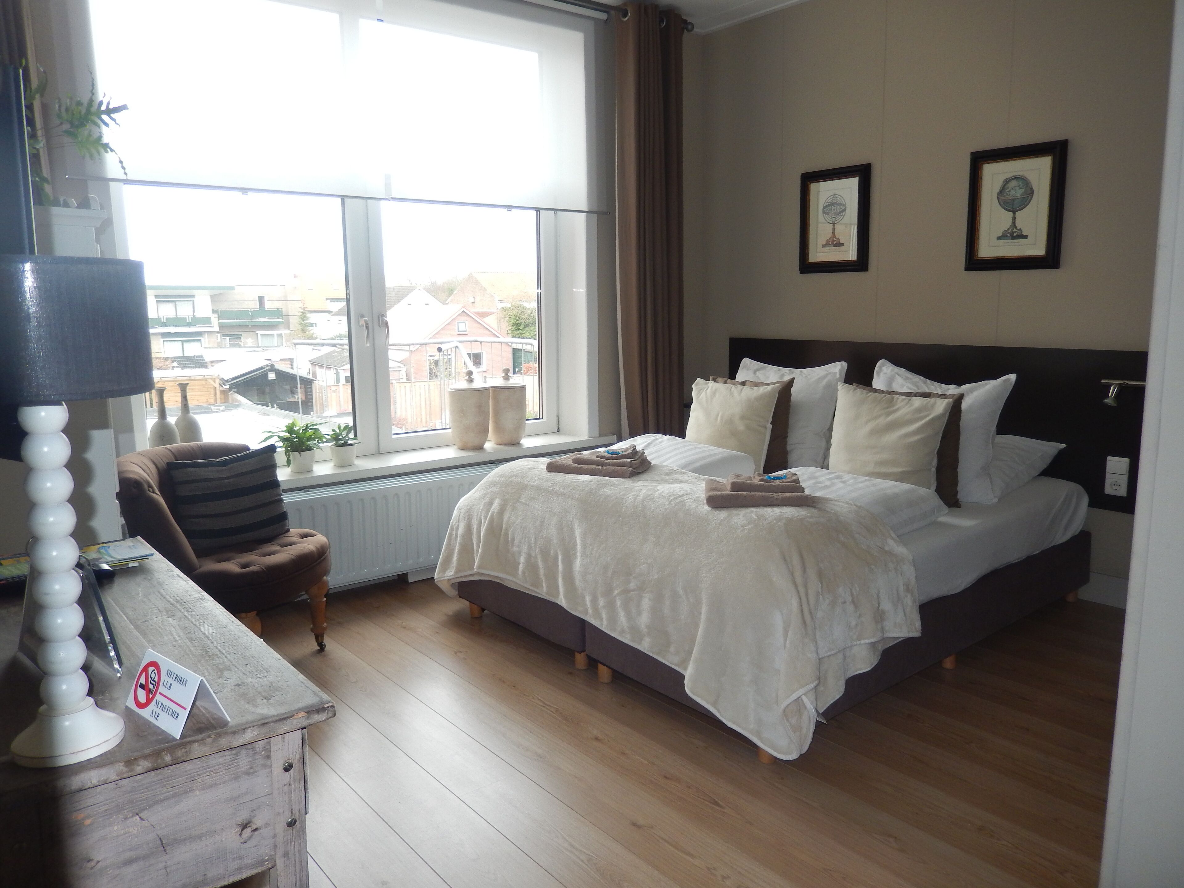 Een slaapkamer met een groot bed, een raam met gordijnen, een nachtkastje met een lamp en twee ingelijste foto's aan de muur.