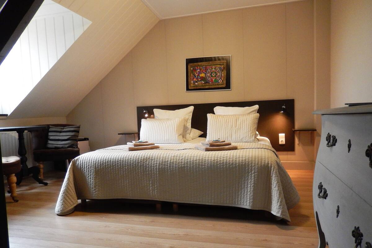 A bedroom with a large bed, a headboard with a clock, a small table with a lamp, and a dresser with decorative handles.