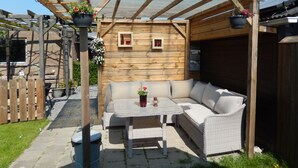 A covered patio area with a wicker sofa and table, wooden pergola, and hanging flower pots. - B&B de Passant (Breskens)