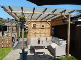 Un espacio de patio cubierto con un sofá y una mesa de mimbre, una pérgola de madera y macetas colgantes con flores.