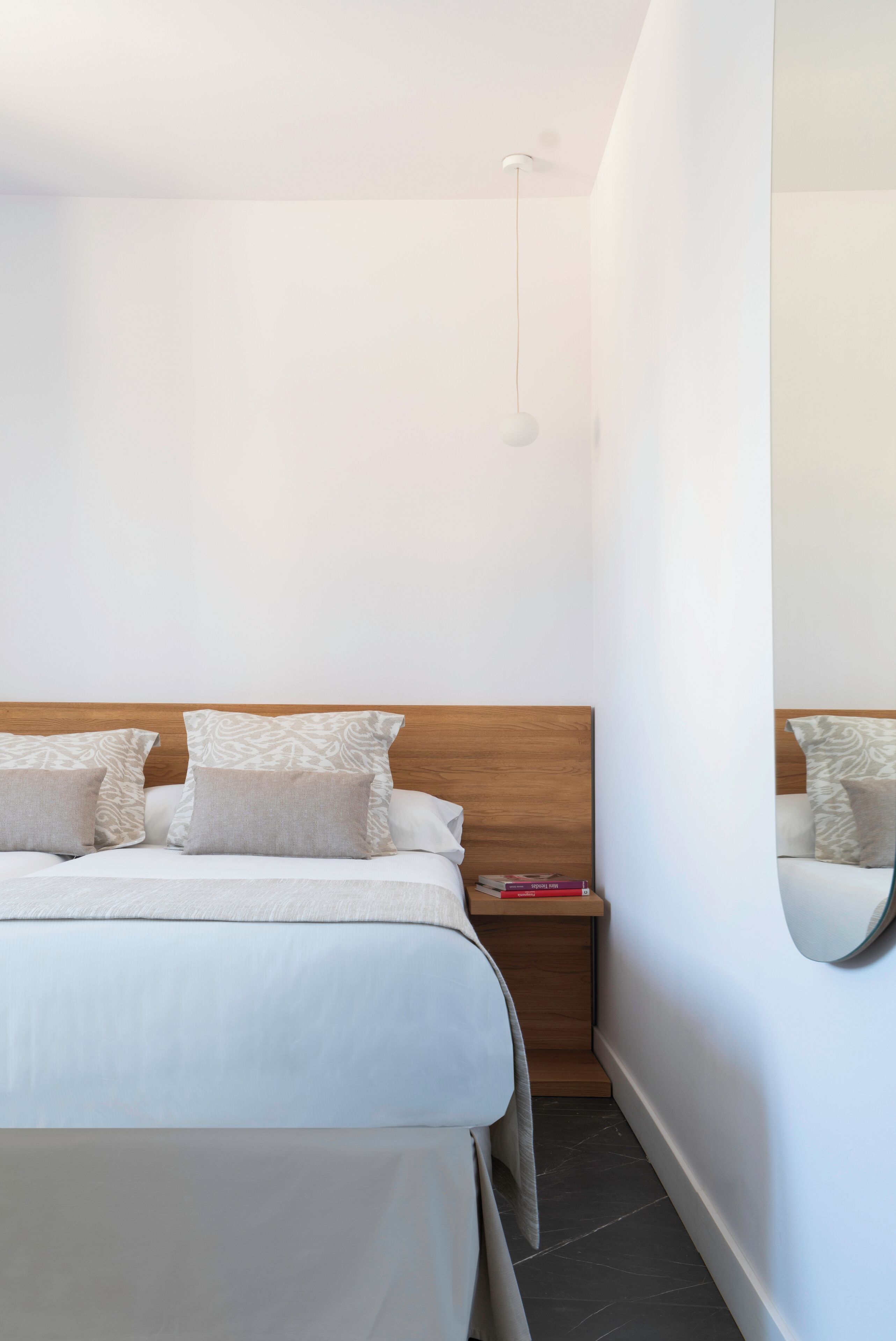 A modern hotel room with a wooden headboard, a bedside table with a red book, and a mirror on the wall.