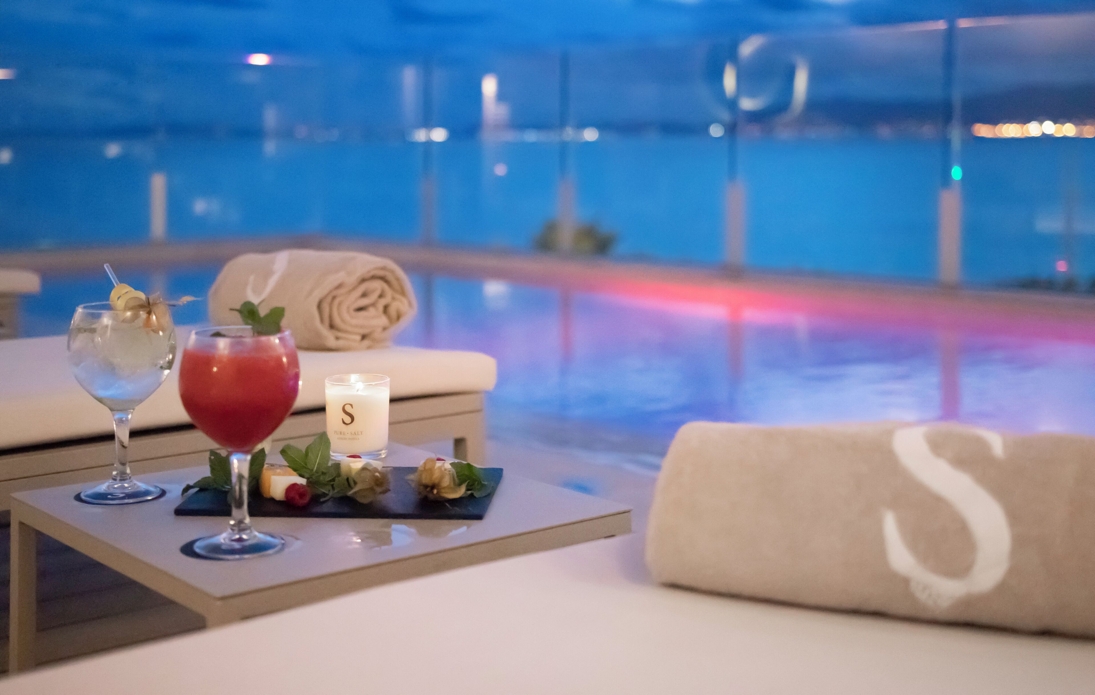 A poolside setting with a table featuring a drink, a candle, and a plate of food.
