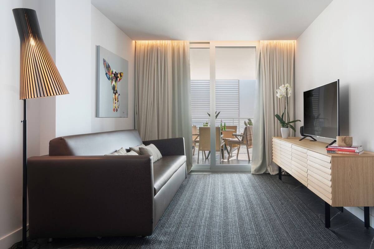 Une chambre d’hôtel moderne, dotée d’un canapé, d’une télévision, d’un bureau agrémenté d’une plante, et offrant une vue sur un balcon à travers une porte vitrée.