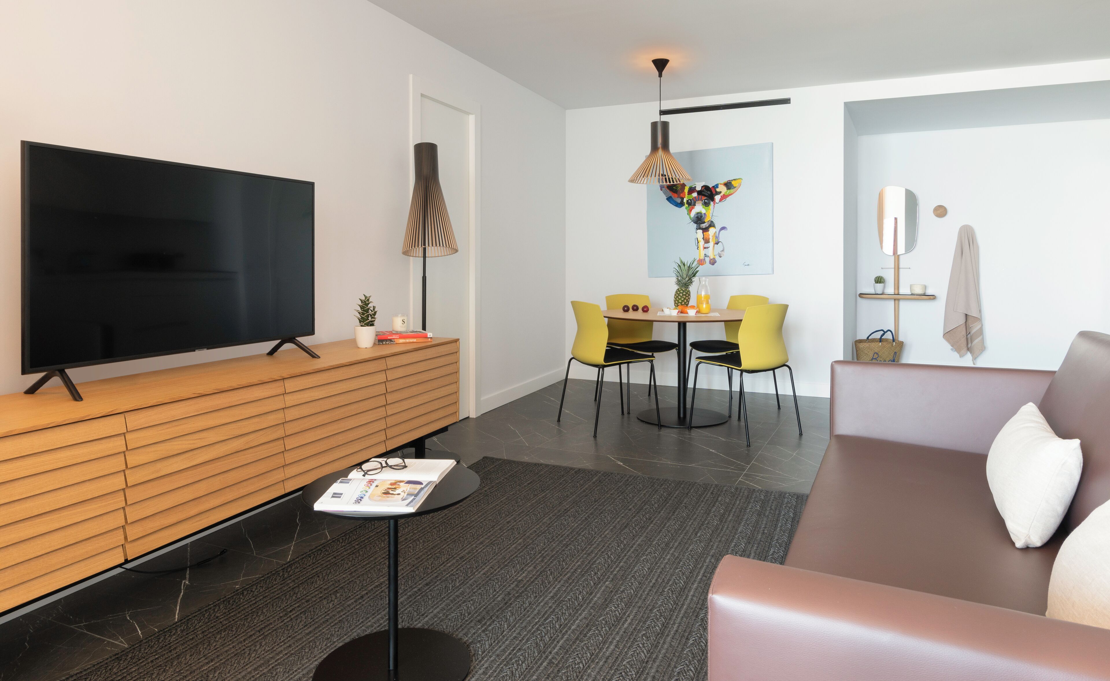 Una sala de estar moderna con un televisor de pantalla plana, una consola de madera, sillas amarillas, un sofá marrón y un comedor con mesa y sillas.
