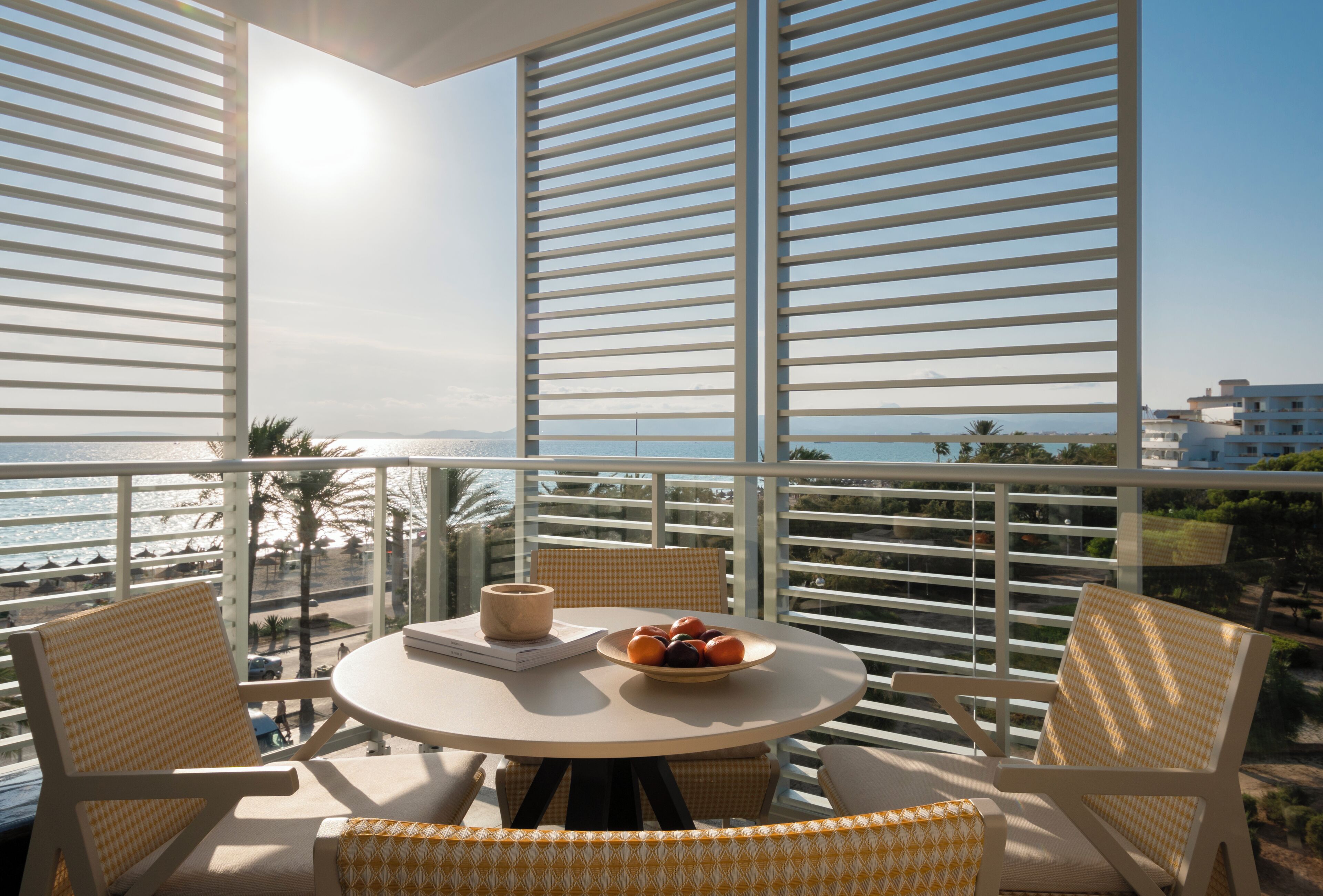 Un balcón con mesa y sillas, un cuenco de fruta y vistas a la playa y edificios.