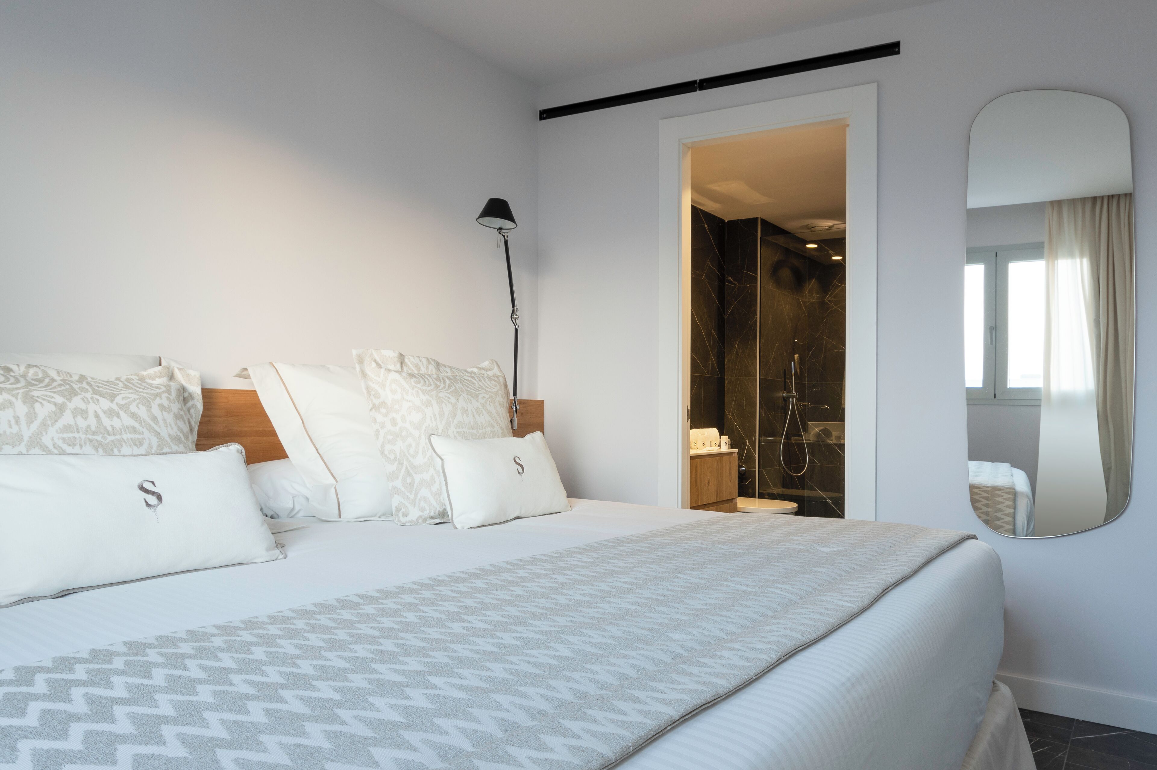A modern hotel room with a large bed, a bedside lamp, a mirror, and a shower area visible through an open door.