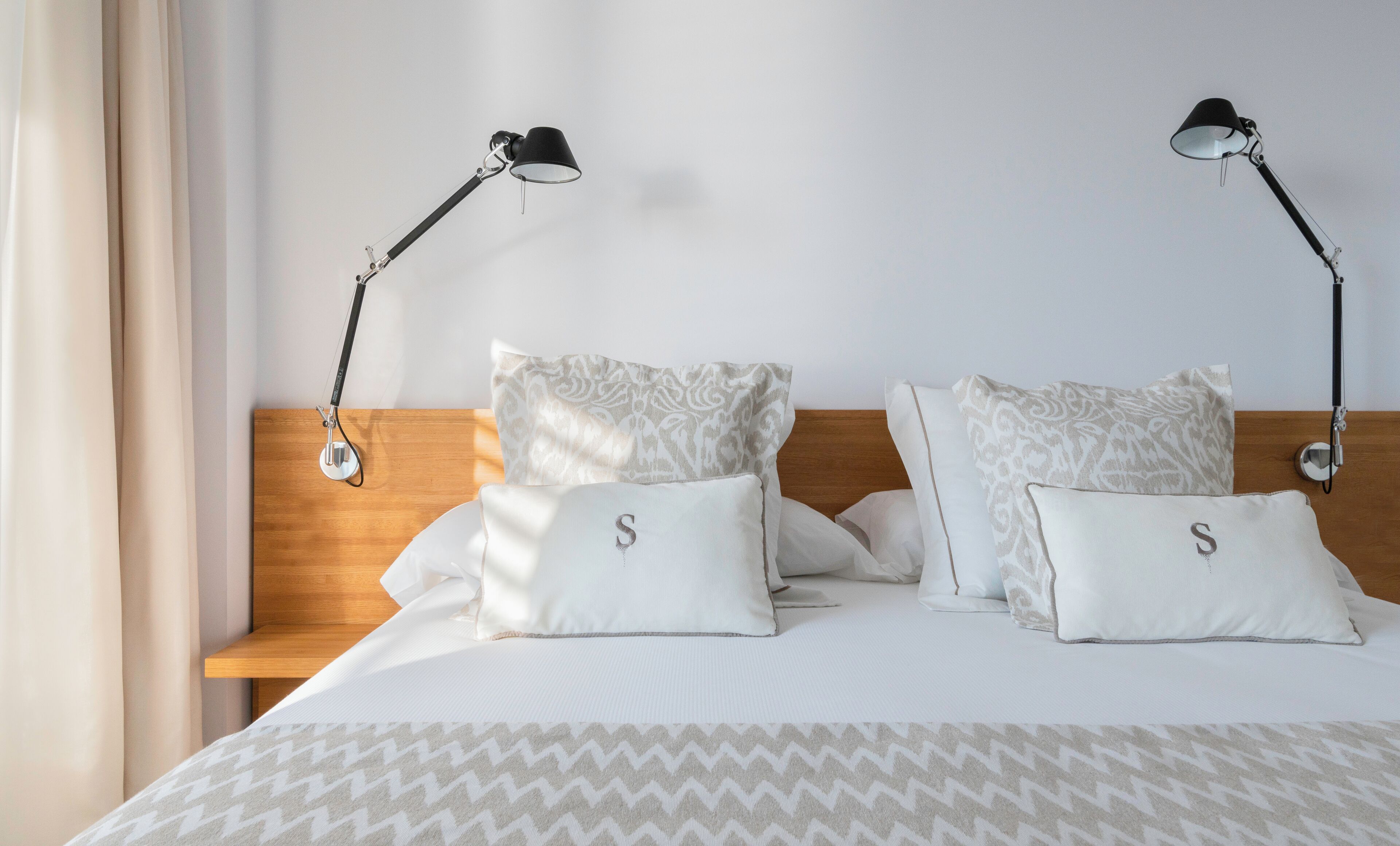 A neatly made bed with two pillows, each with a 'S' on them, and two bedside lamps on a wooden headboard.