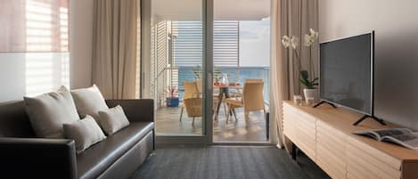 Quarto de hotel moderno com sofá, televisão, mesa lateral de madeira e varanda com vista para o mar.
