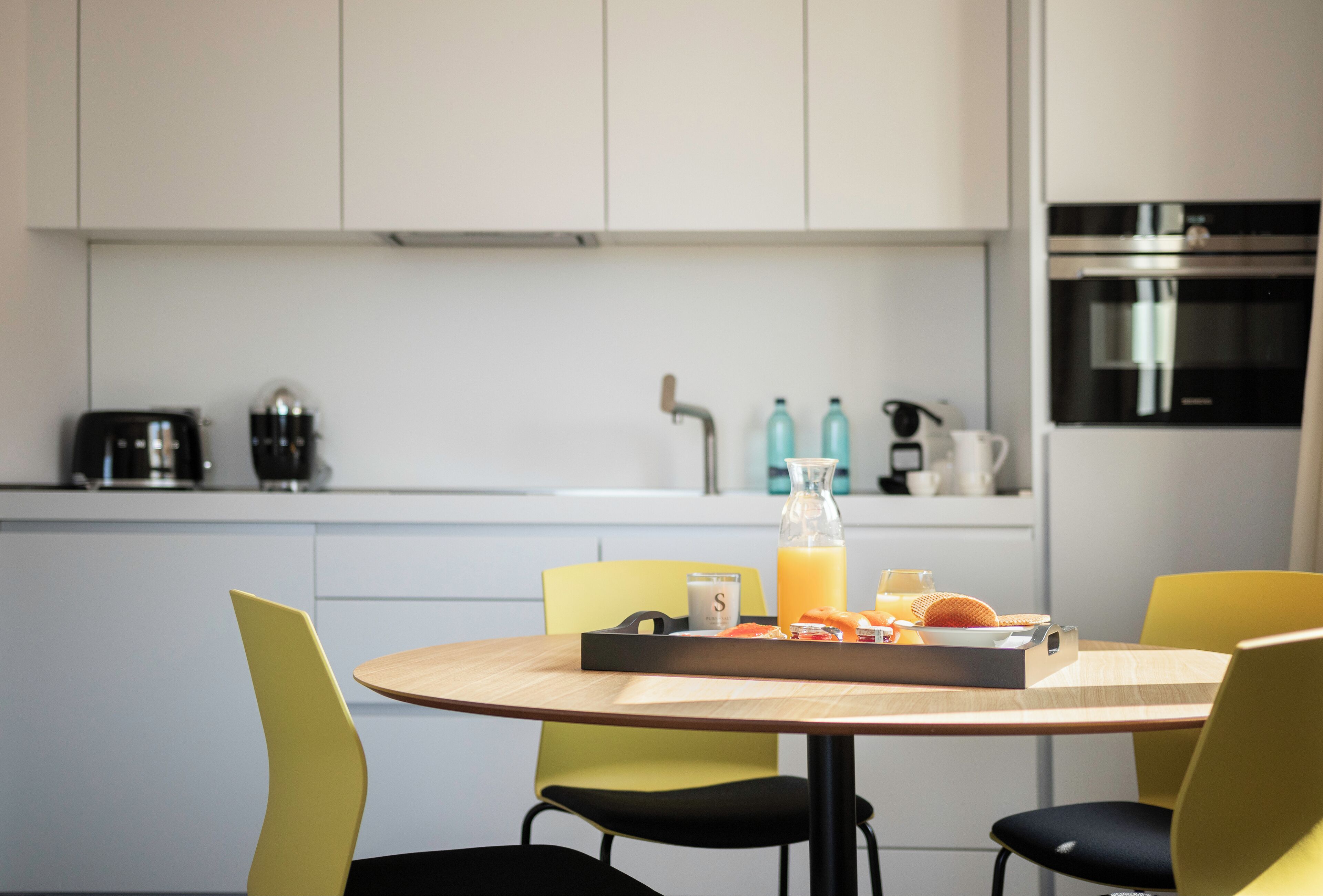 A modern kitchen with white cabinets, a stainless steel oven, and a dining area with yellow chairs and a wooden table.