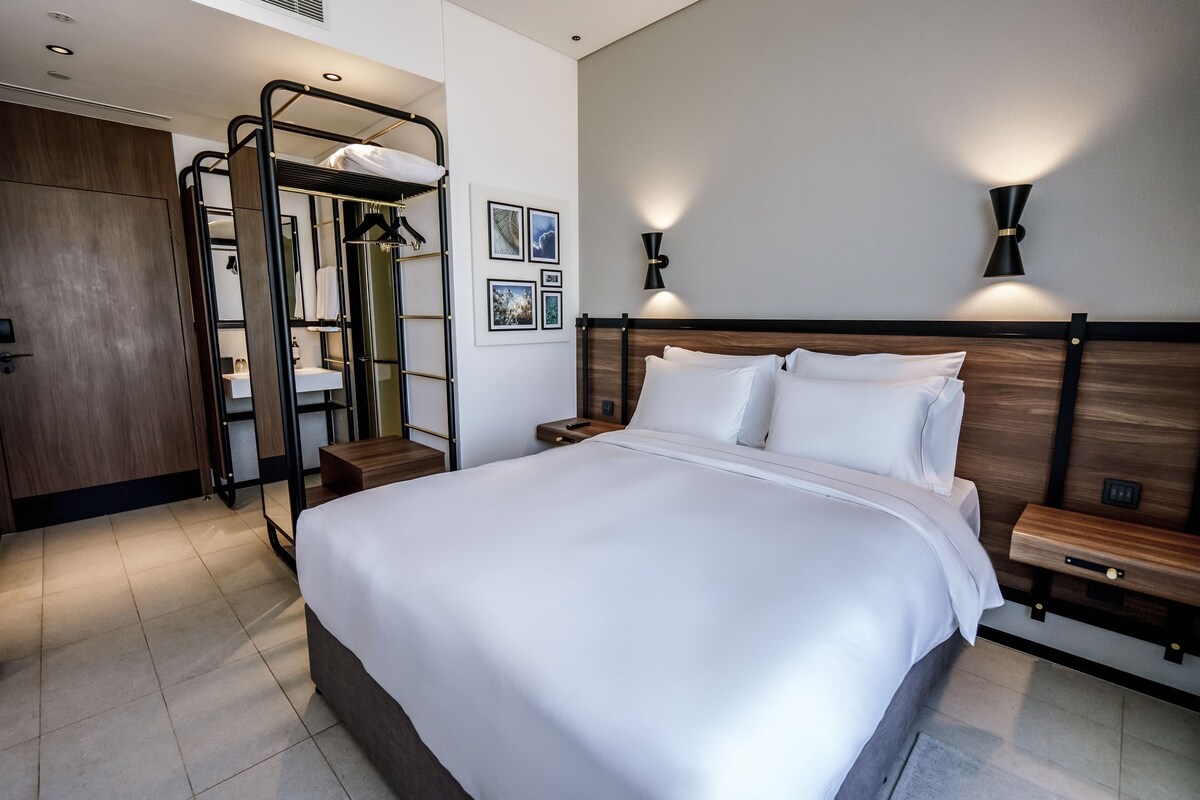 A hotel room with a large bed, a wooden wardrobe, a mirror, a small bathroom, and two bedside tables.