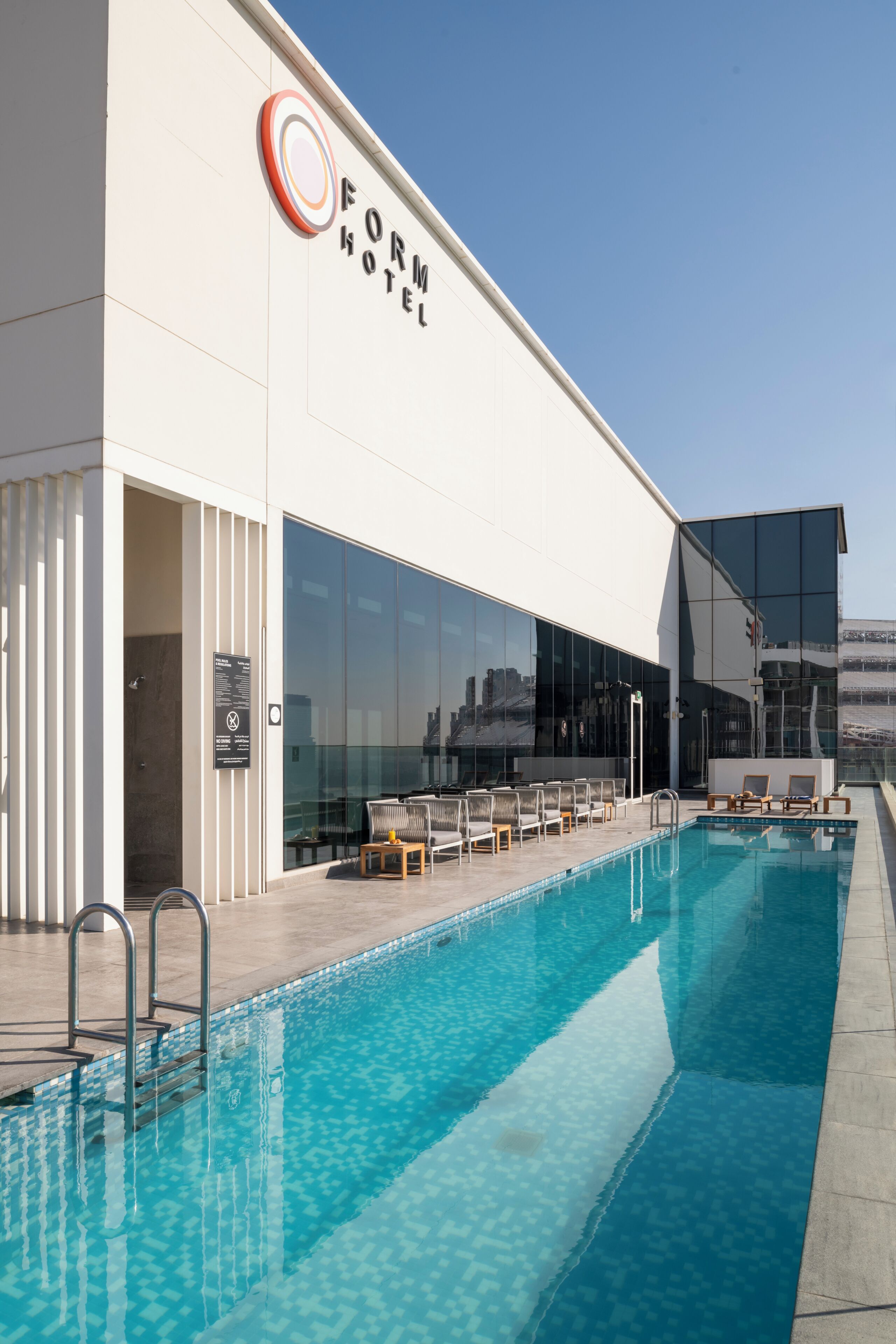 A modern hotel with a swimming pool, outdoor seating, and a clear sky.