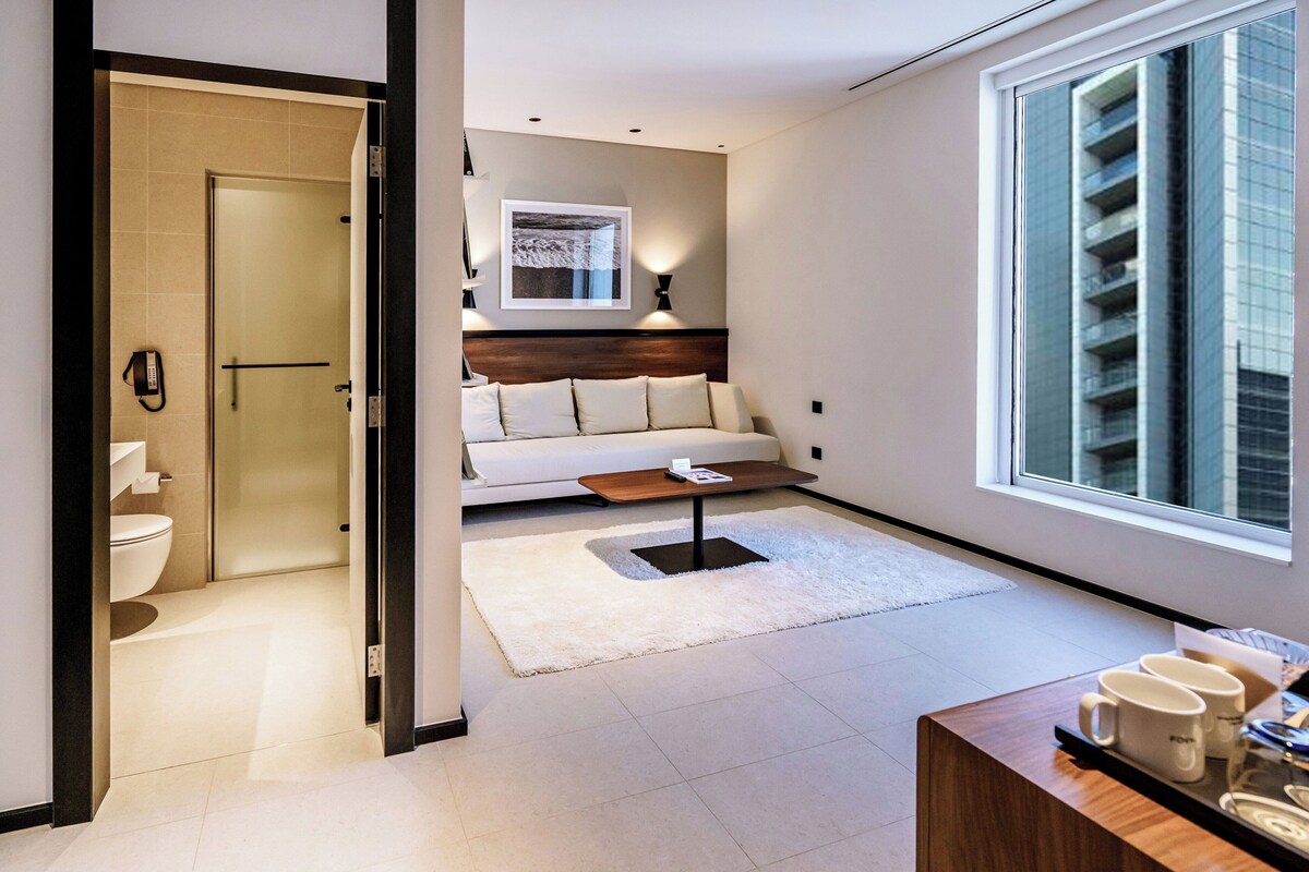 A modern hotel room with a glass door leading to a bathroom, a sofa with a wooden coffee table, and a large window with a city view.