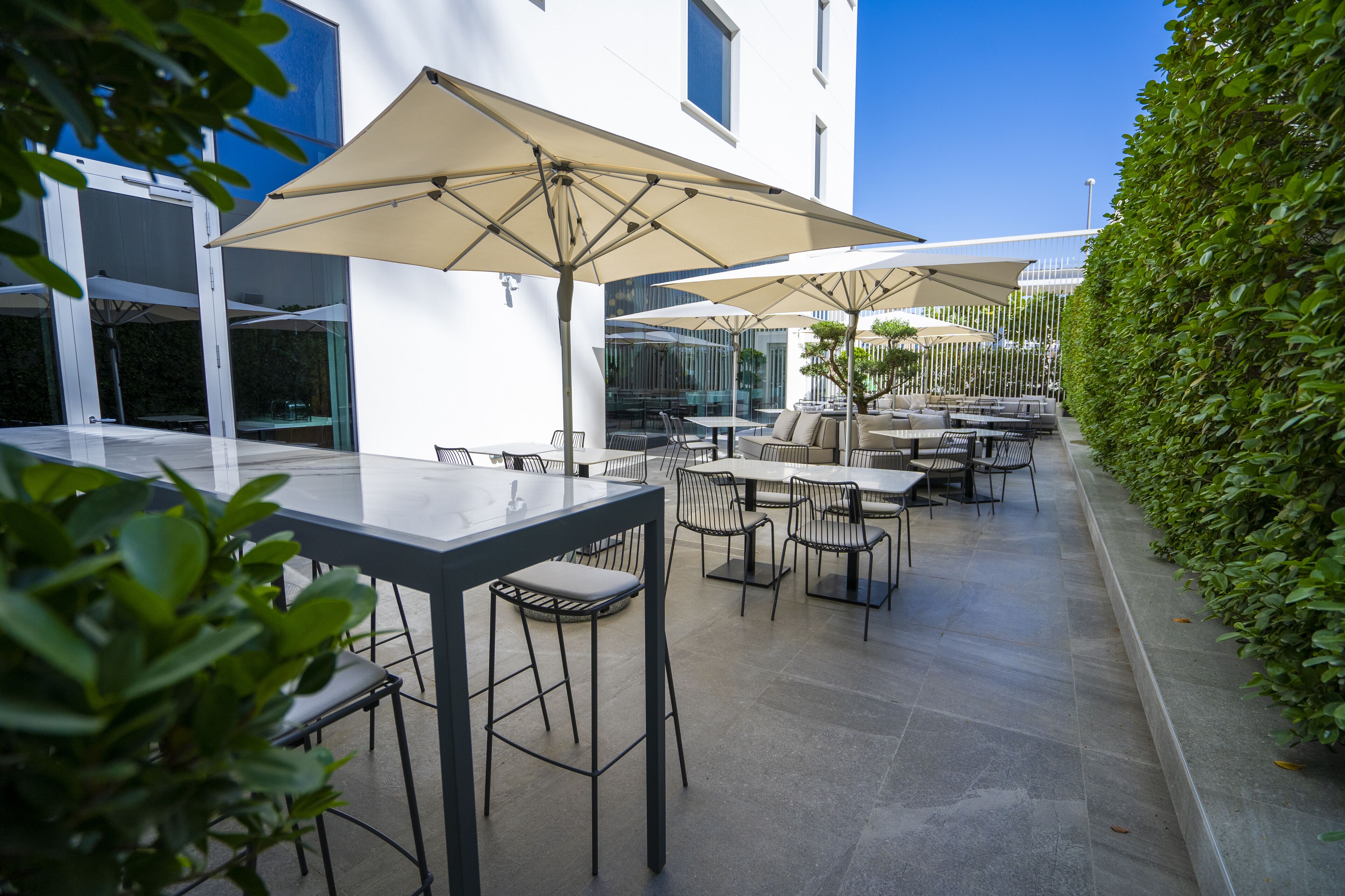 Un patio exterior con mesas y sillas, sombrillas grandes y un seto a lo largo de un lado.