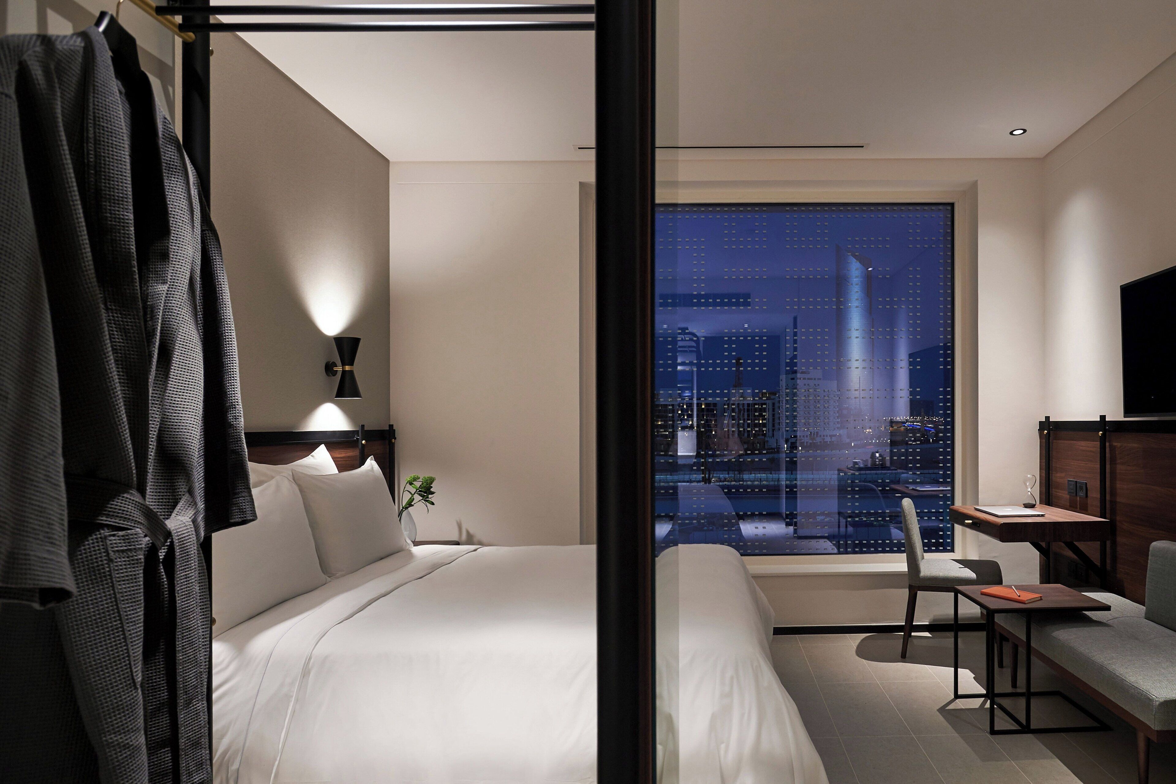 A modern hotel room with a large bed, a desk, a chair, and a view of a cityscape at night.