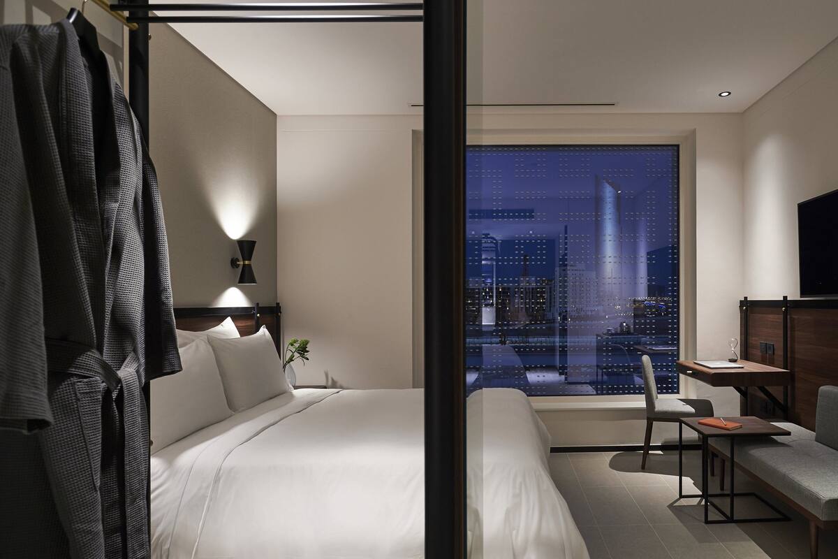 A modern hotel room with a large bed, a desk, and a chair. There is a view of a cityscape through a large window.