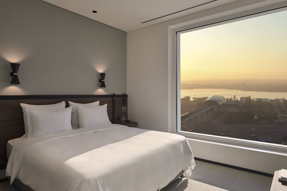 A modern hotel room with a large bed, two wall-mounted lights, and a large window offering a city view.