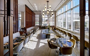 A modern restaurant with long communal tables, individual seating, and large windows offering city views.