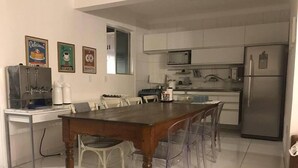 A modern kitchen with white cabinets, a stainless steel refrigerator, and a wooden dining table with clear plastic chairs. - Hostel Pajuçara (Maceió)