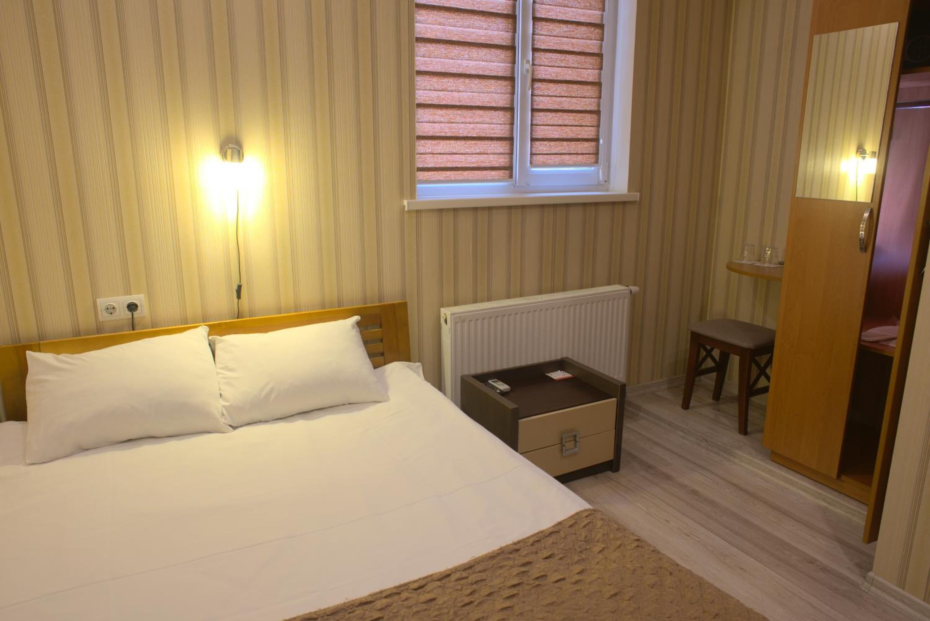 Une chambre à coucher comprenant un lit, une table de chevet, une commode, un radiateur, une fenêtre avec stores et une armoire.