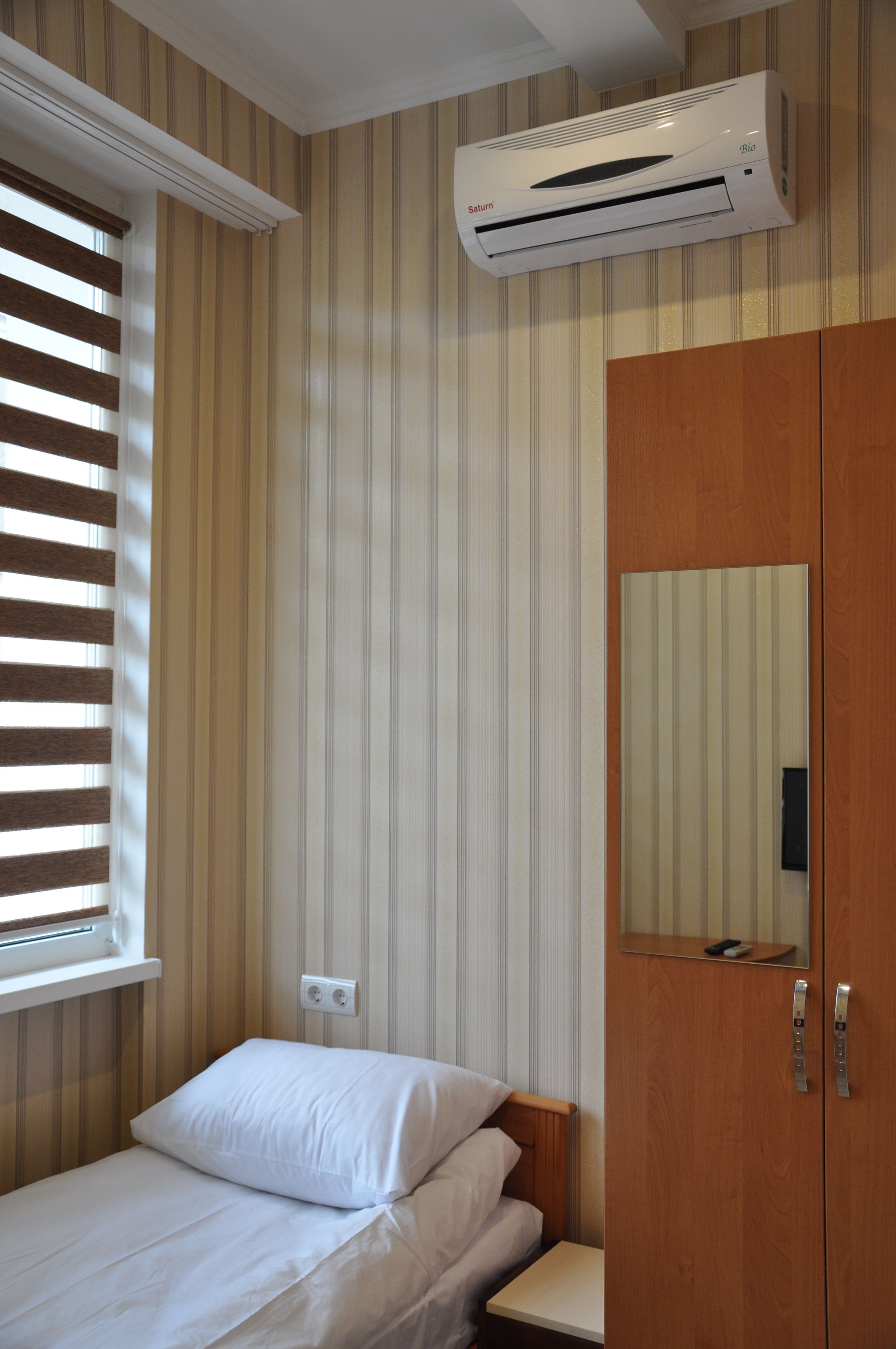 A bedroom with a bed, a wardrobe, a window with blinds, and an air conditioning unit.