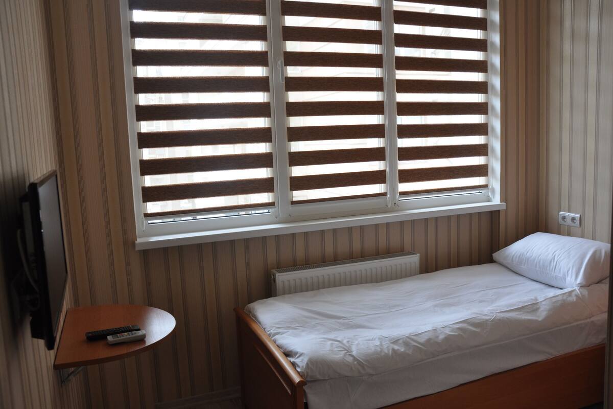 A small, single-bed room with a wooden headboard, a bedside table with a remote control, and a window with blinds.
