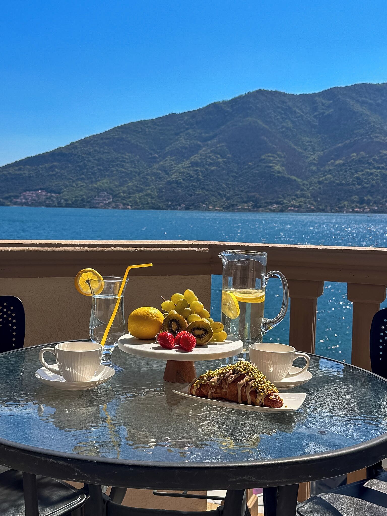 A table with a glass pitcher, cups, fruits, and a pastry on a balcony overlooking a lake and mountains.