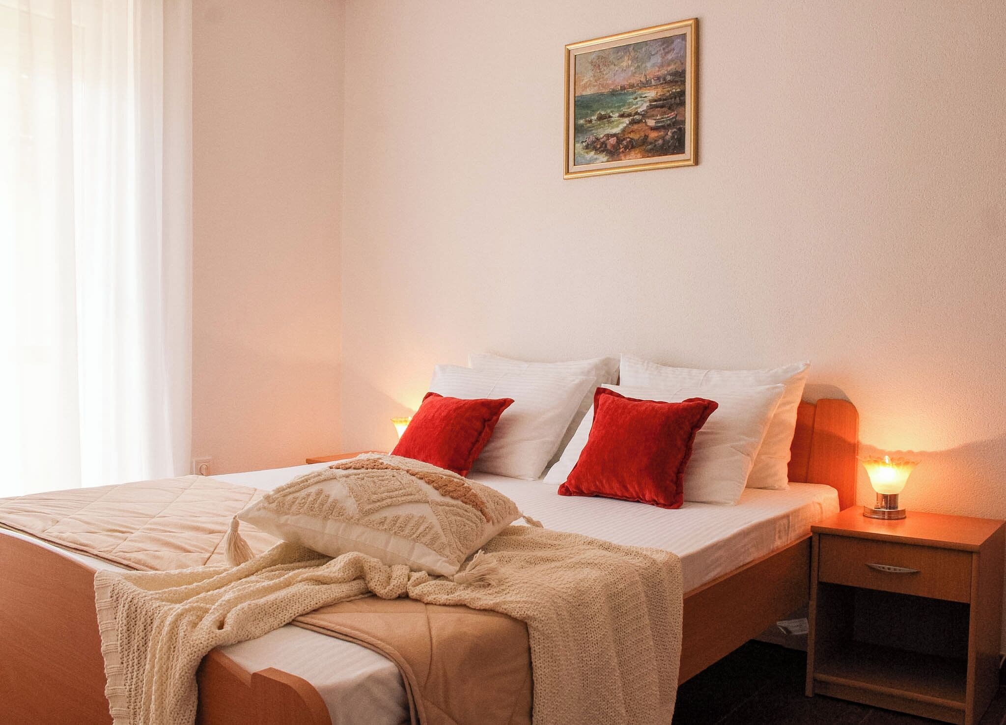 A hotel room with a bed, pillows, a blanket, a nightstand, a lamp, and a painting on the wall.