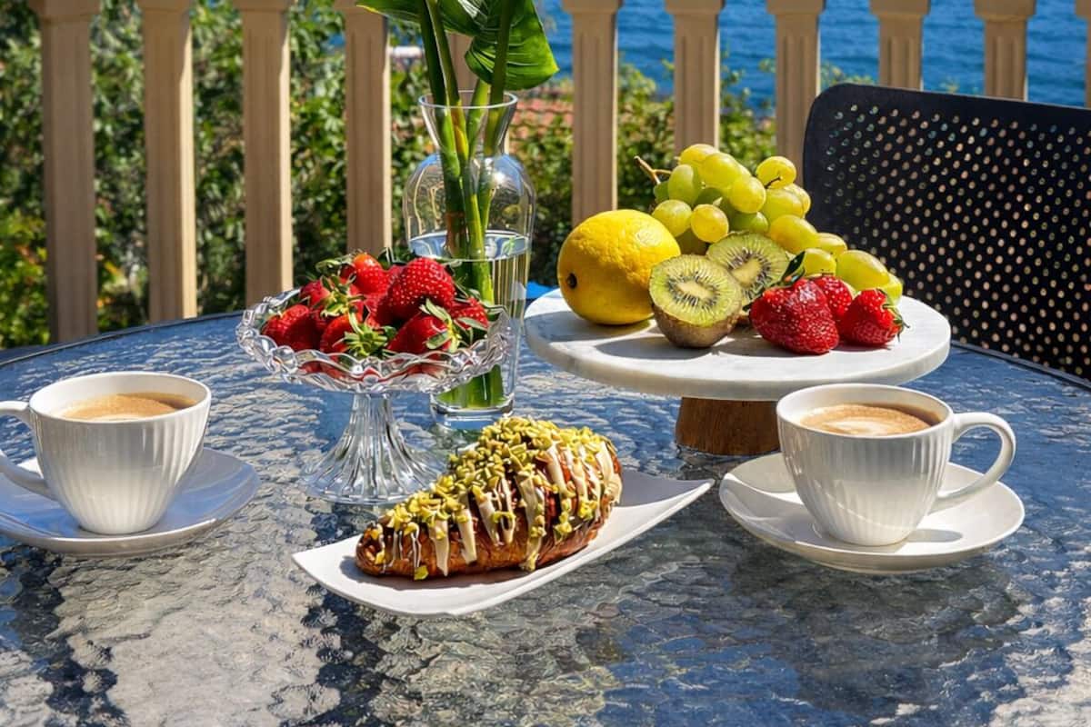 Une table avec vue sur un lac et des montagnes, un vase rempli de fleurs blanches, un panier de fruits et des pâtisseries.