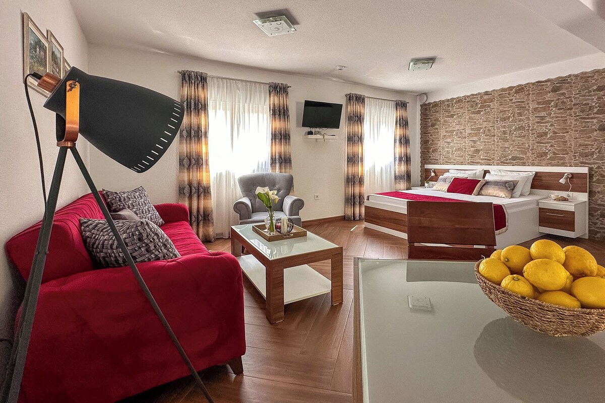 A room with a red sofa, a wooden coffee table, a grey armchair, a bed with a stone wall, and a television on the wall.