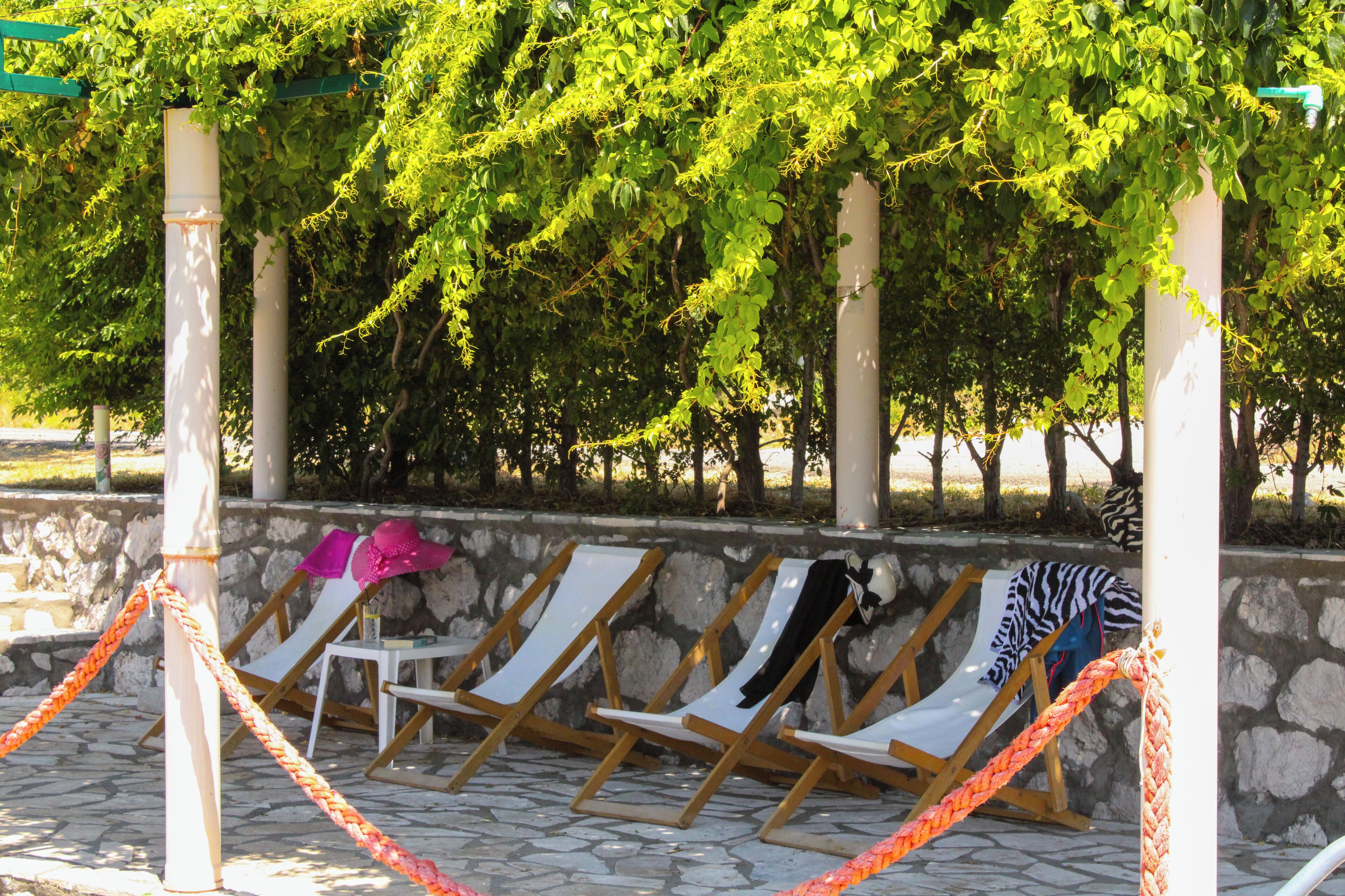 A row of sun loungers under a pergola with a stone wall backdrop.