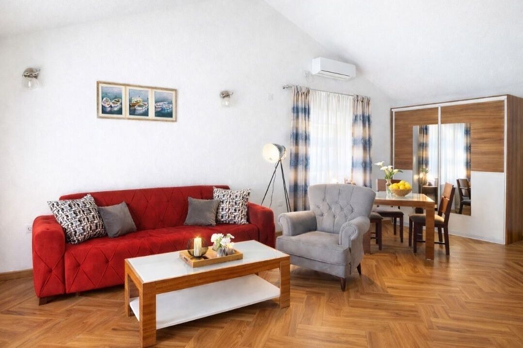 Een woonkamer met een rode bank, een grijze fauteuil, een houten salontafel, en een eethoek met een tafel en stoelen.