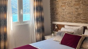 A bedroom with a stone wall, a bed with a red blanket, a nightstand, a lamp, and a window with curtains. - Apartments Stella del Mare (Risan)