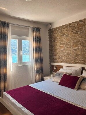 Un dormitorio con pared de piedra, cama con manta roja, mesita de noche, lámpara y ventana con cortinas.