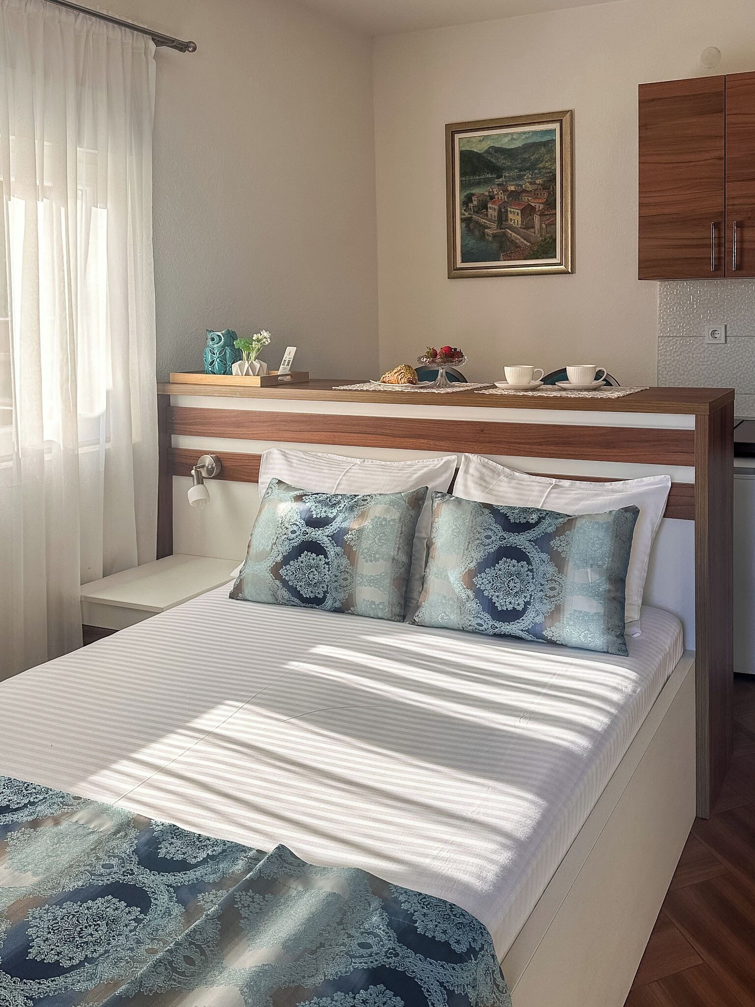 A bedroom with a bed, a bedside table, a nightstand, a window with sheer curtains, a painting on the wall, and a kitchenette area.